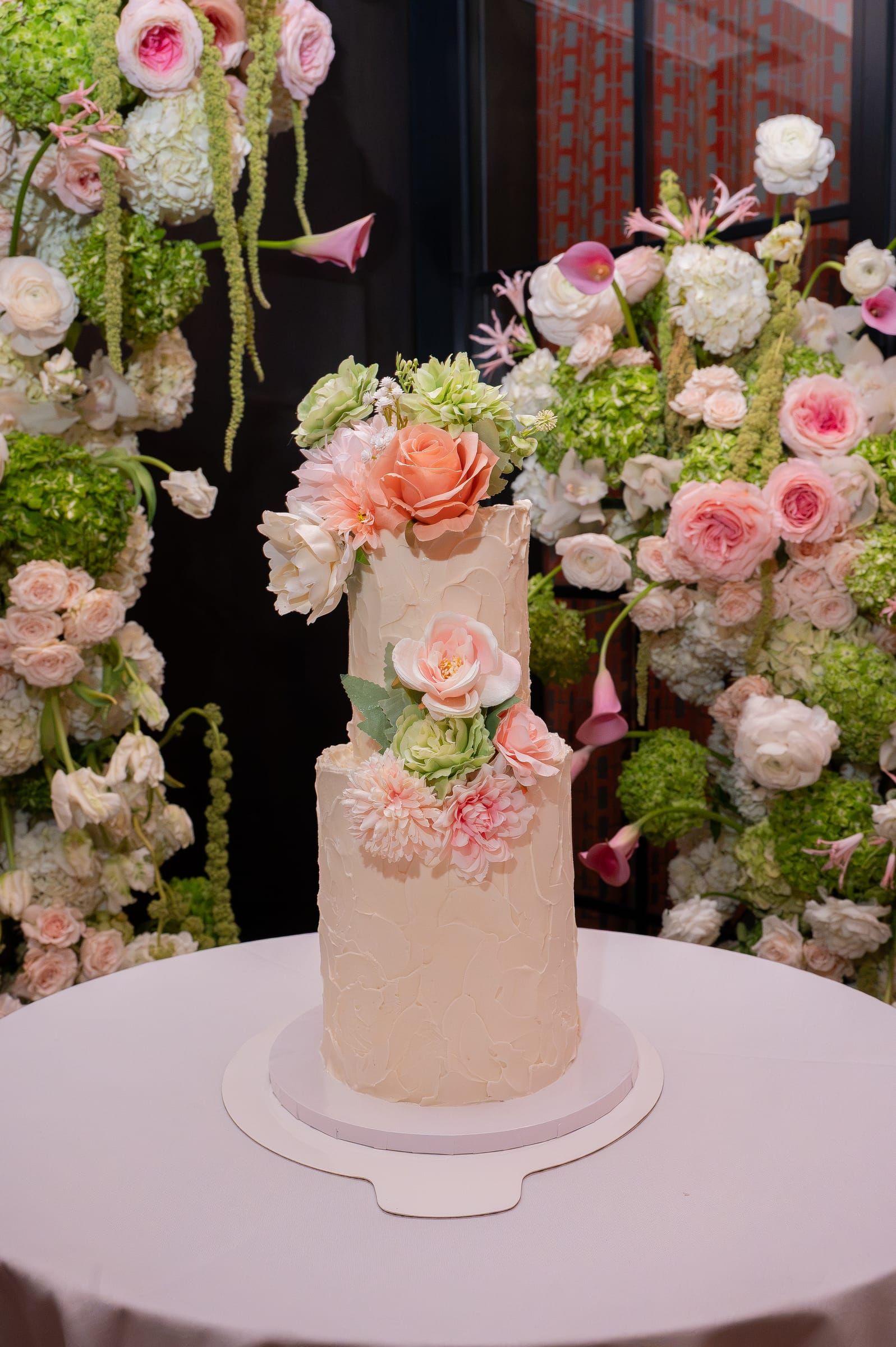 A two-tier white cake topped with peach and blush sugar flowers, in front of a floral wall.