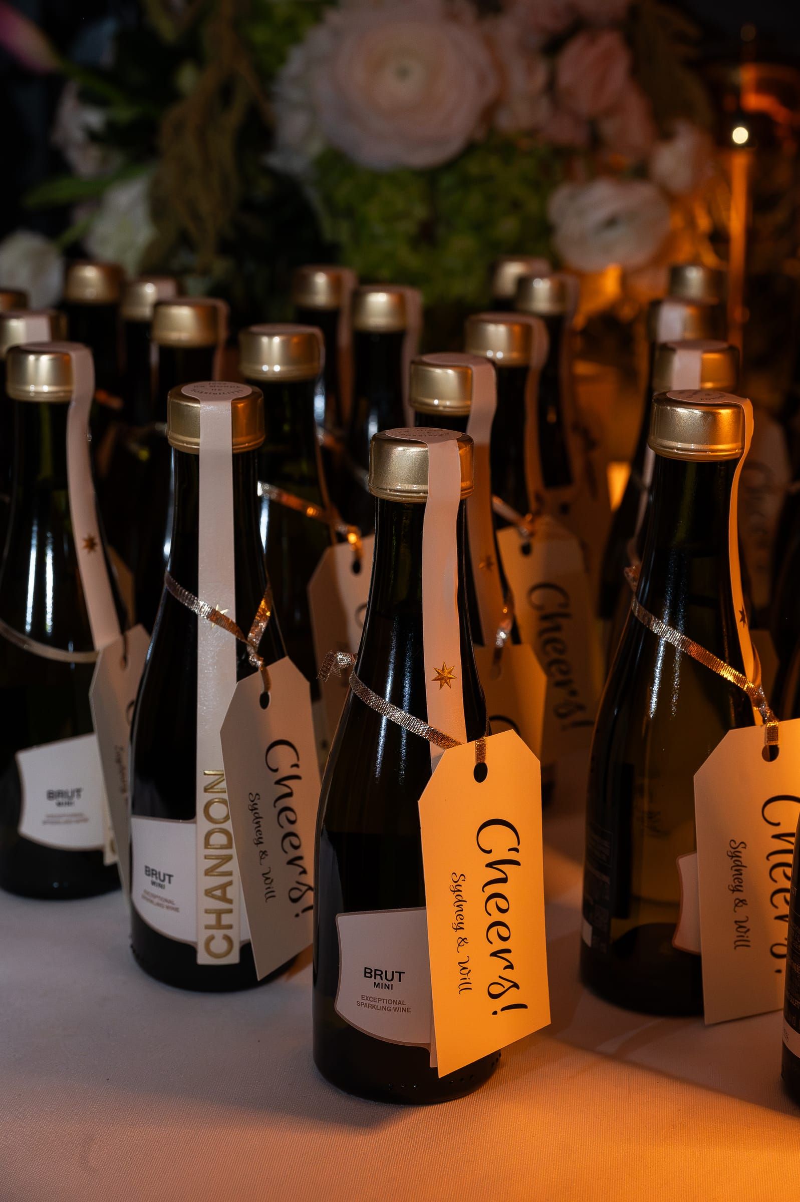 Mini bottles of champagne tied with handwritten “Cheers!” tags, set out as favors.