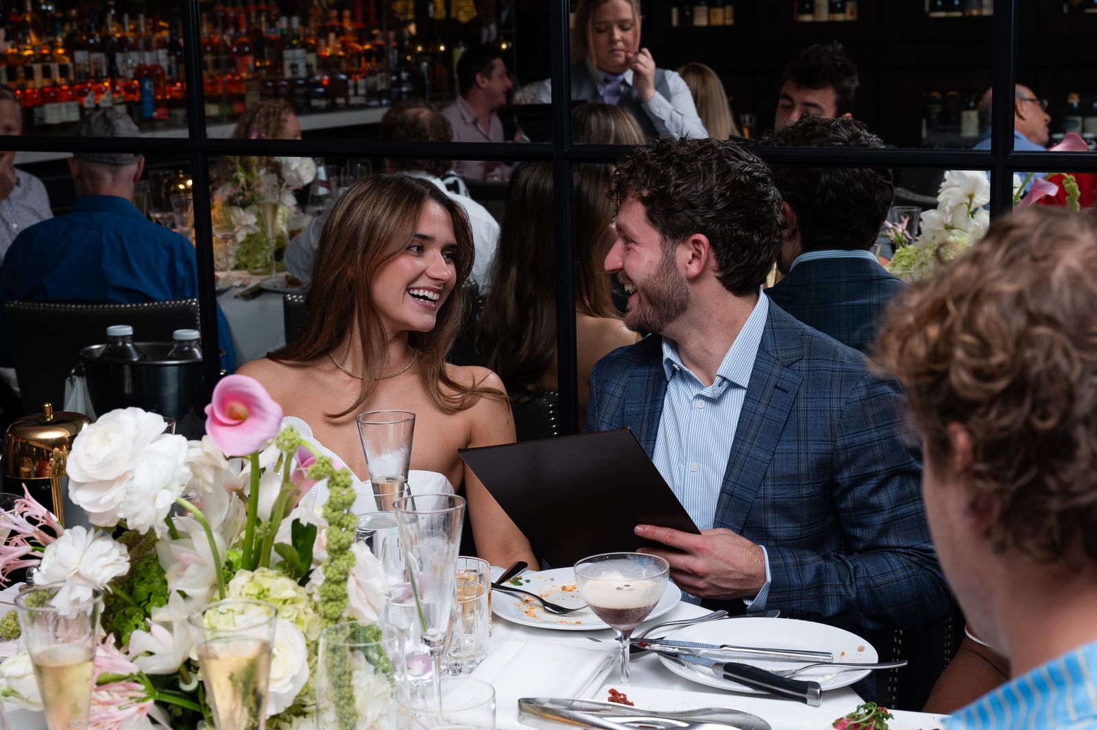 The engaged couple laughing together at the dinner table behind white florals.
