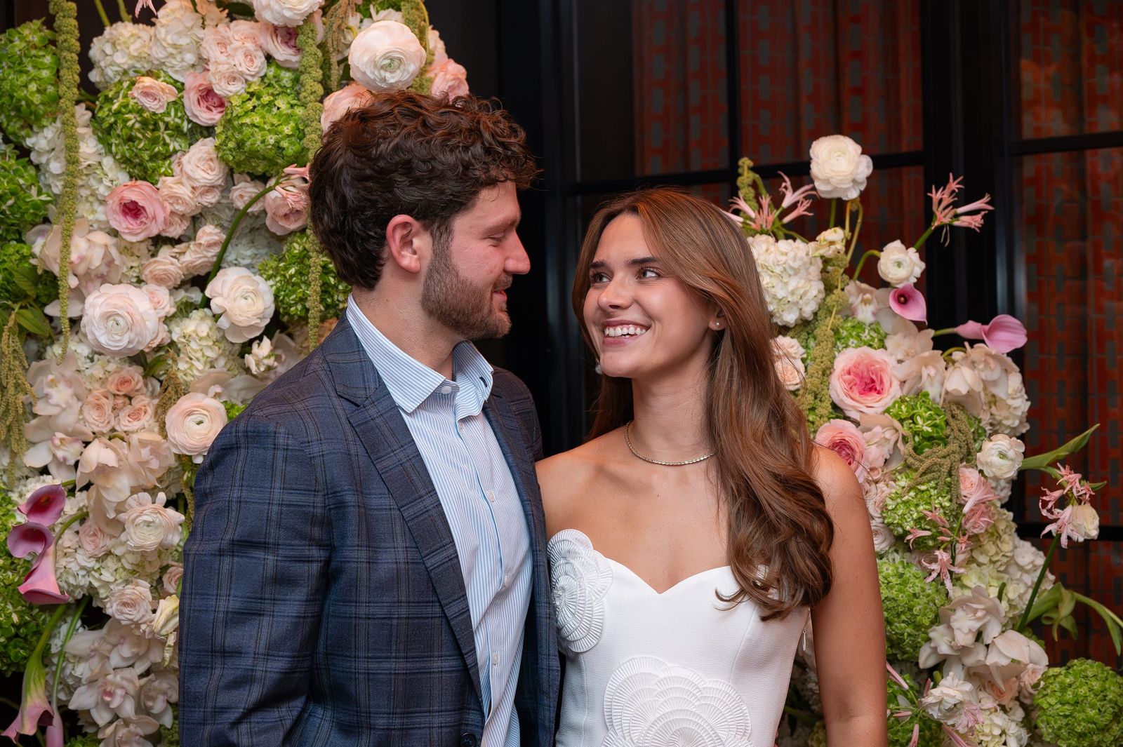 The engaged couple smiling at each other in front of a wall of roses, hydrangea and calla lilies.