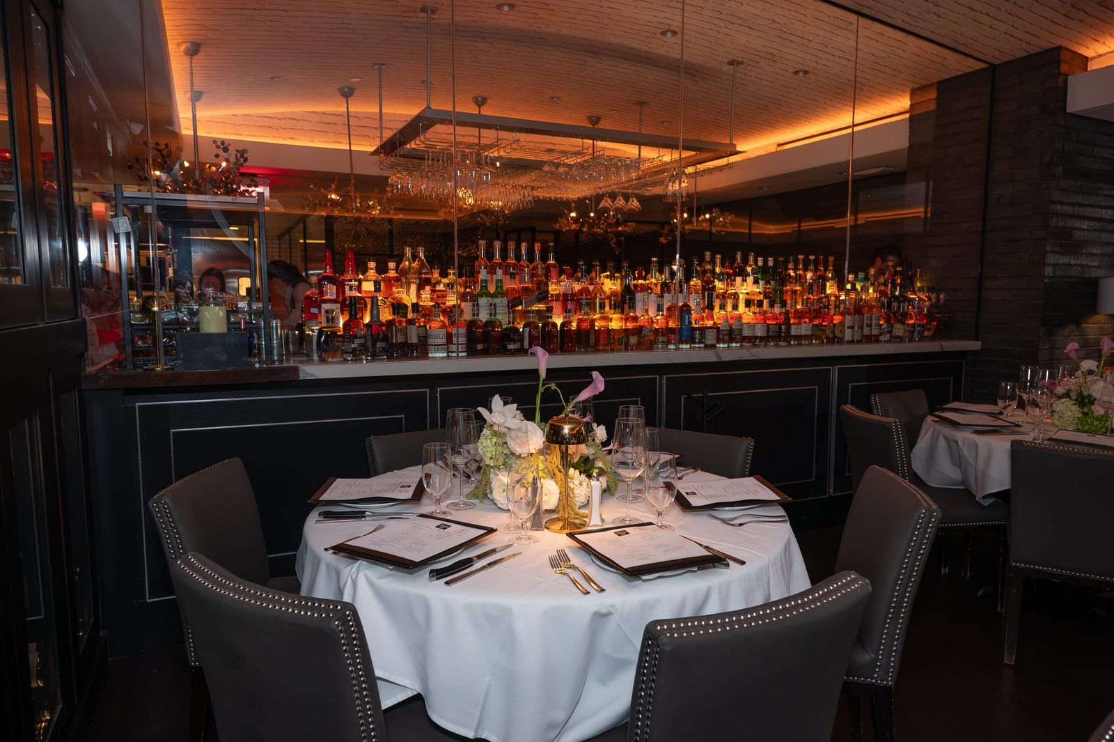 A round table dressed in white linen and pink and green florals, in a dining room glowing amber in front of a backlit bar.