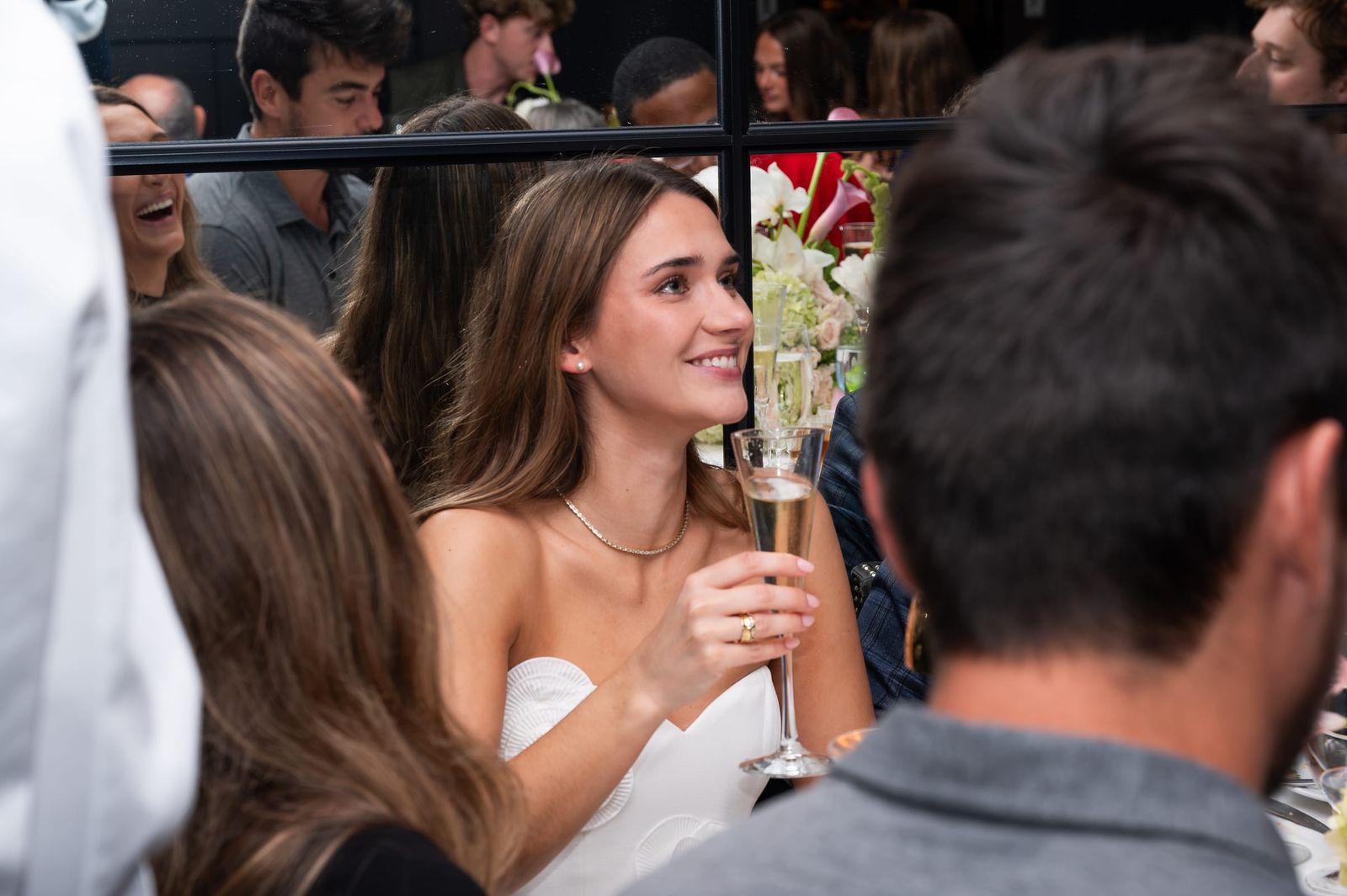 The guest of honor smiling with a glass of champagne as friends gather around her.