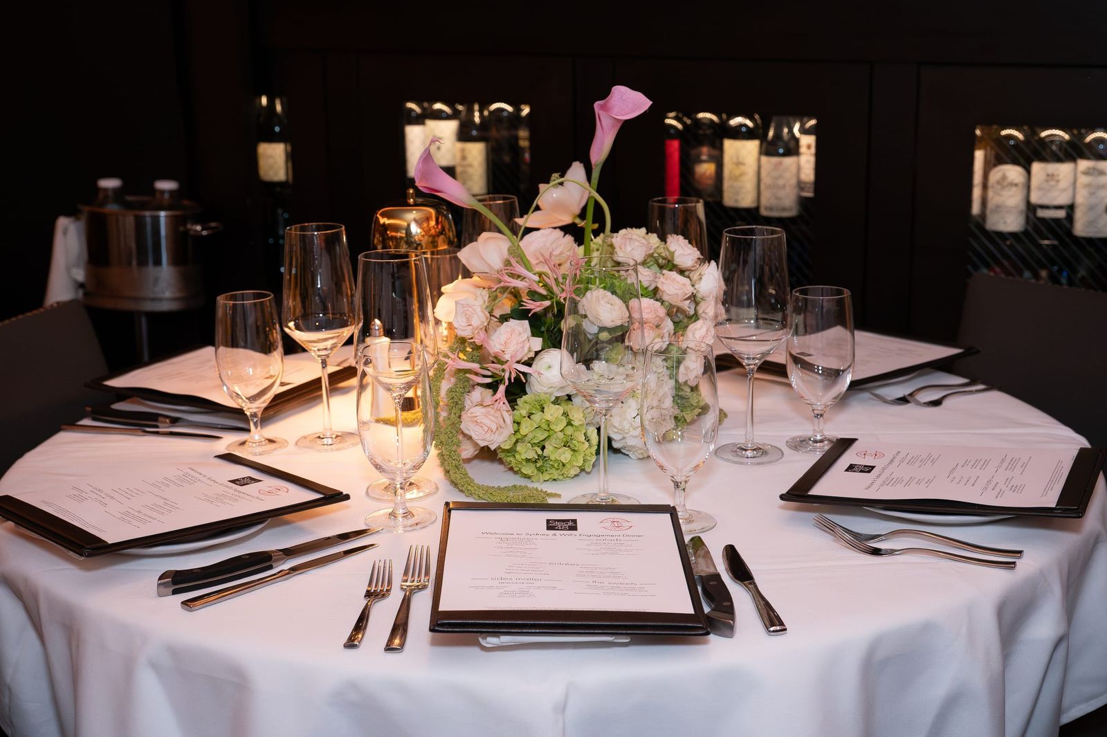 A tablescape of blush roses, green hydrangea and pink calla lilies on white linen, a printed menu at every place.