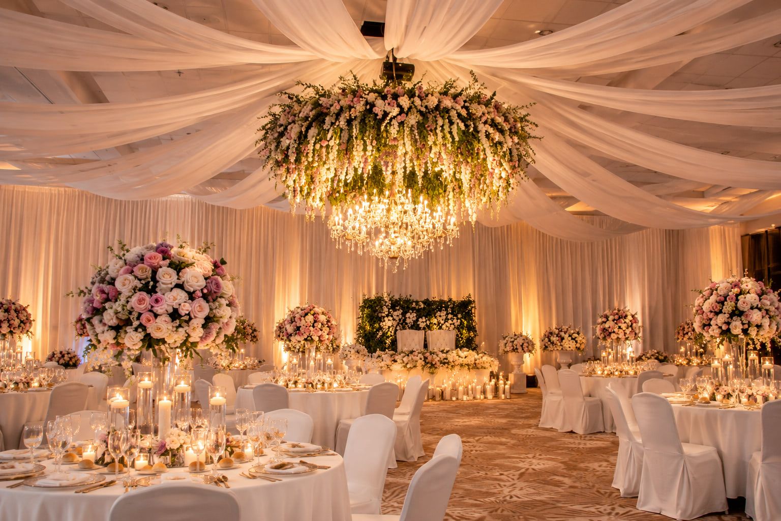 A ballroom hung with ivory drapery and a floral chandelier above candlelit round tables.